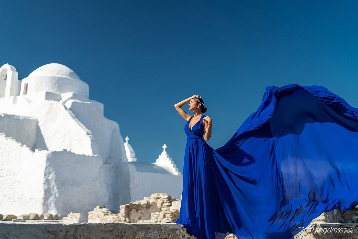 Woman in a royal blue flying dress posing in front of a white-washed church in Mykonos, Greece, with the dress dramatically flowing in the wind against a deep blue sky.